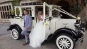 Bride and groom posing next to a cream vintage Imperial wedding car in classic 1930s styling.