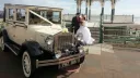 Bride and groom standing beside a cream 1930s style Imperial vintage wedding car with chauffeur and period details.