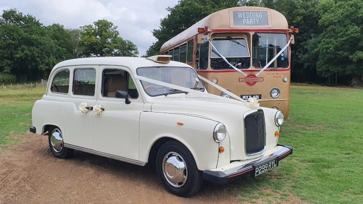 Cream classic London taxi wedding car parked on grass with a vintage gold single-decker bus in the background.