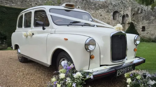 Front three-quarter view of a cream classic London taxi cab decorated for wedding hire with floral details.