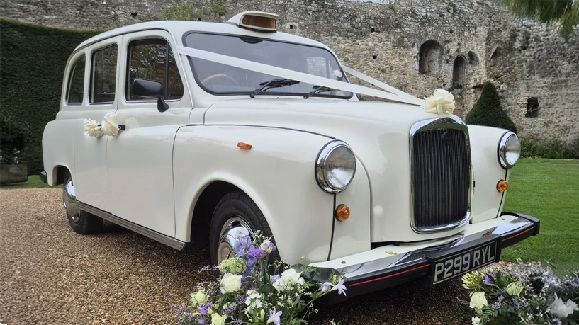 Front three-quarter view of a cream classic London taxi cab decorated for wedding hire with floral details.
