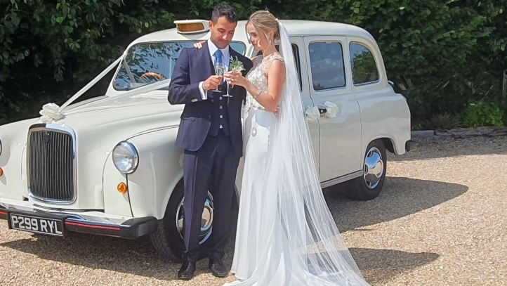 Bride and groom standing next to a cream classic London taxi wedding car outside the venue.