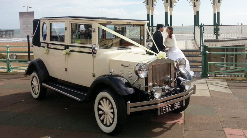 Cream 1930s style Imperial vintage wedding car parked at a seafront location with bride and groom beside the vehicle.