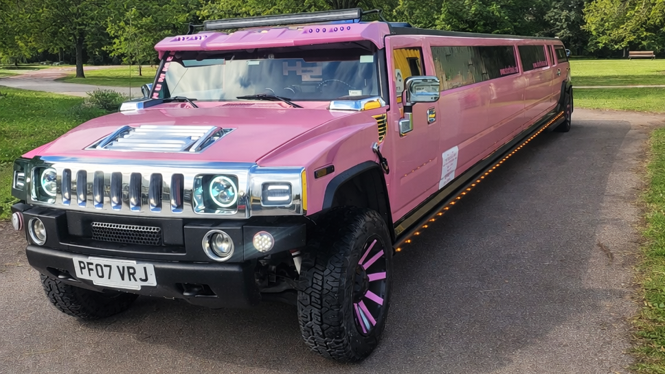 Pink Hummer stretch limousine parked outdoors, front angle view, chrome grille, large pink alloy wheels, party limo for hire.