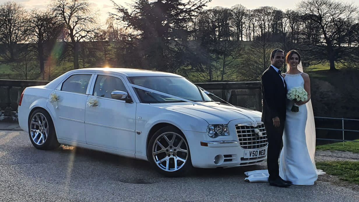 Bride and groom standing beside white Chrysler 300C wedding car with chrome wheels at sunset