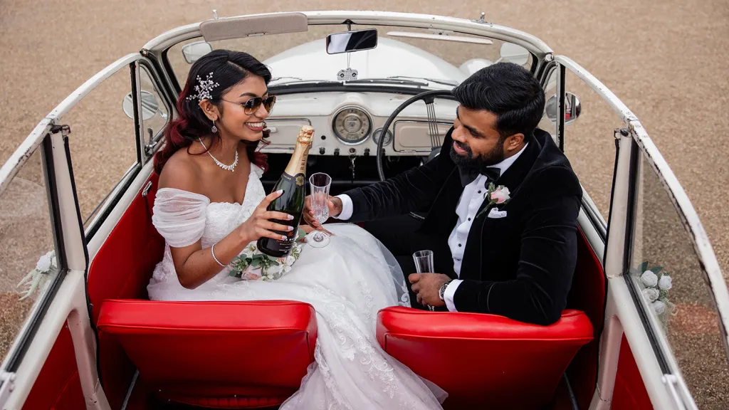 Bride and groom seated inside cream Morris Minor Convertible with bright red leather seats having a celebration toast