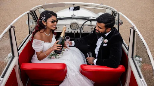 Bride and groom seated inside cream Morris Minor Convertible with bright red leather seats having a celebration toast