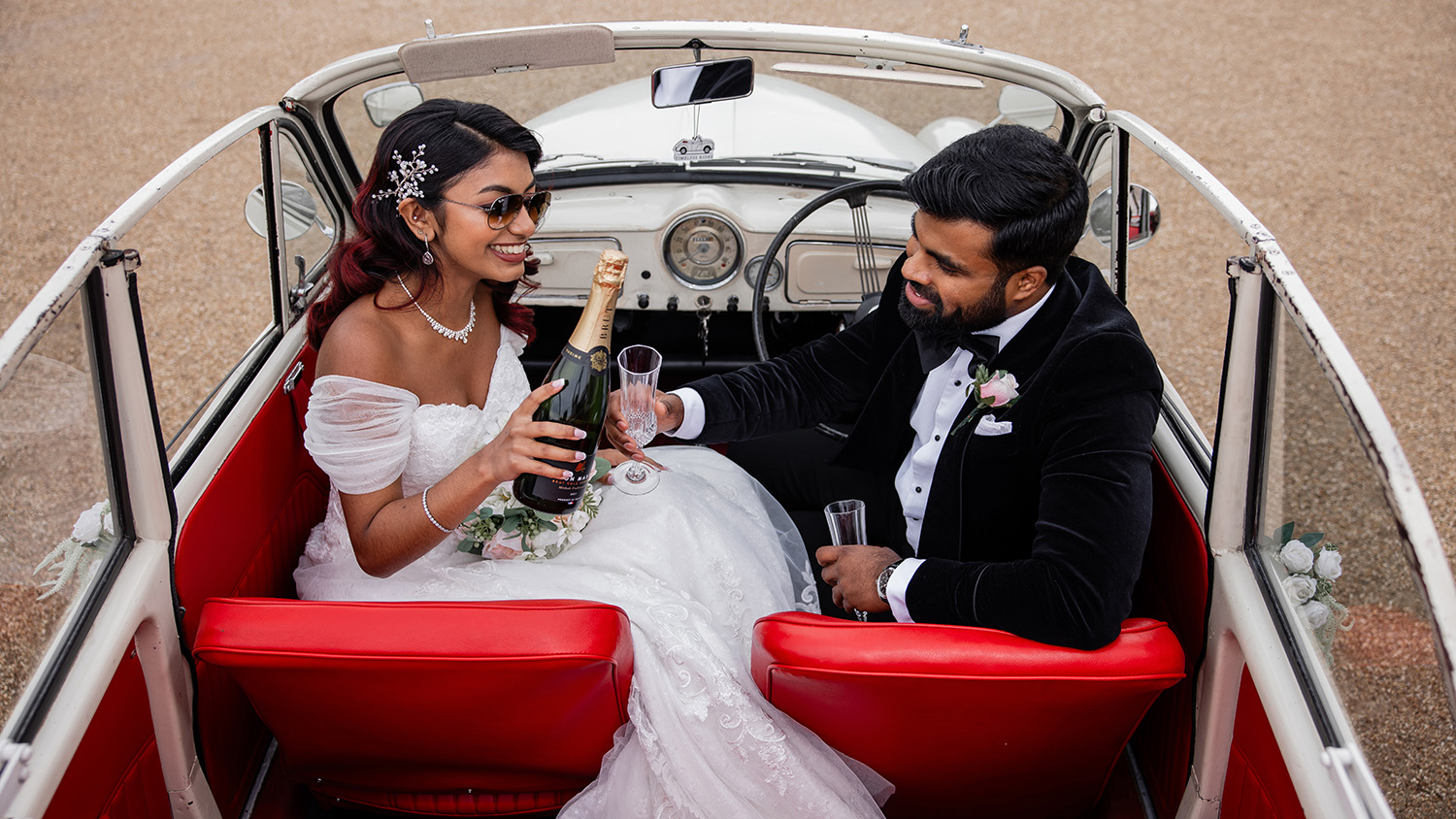 Bride and groom seated inside cream Morris Minor Convertible with bright red leather seats having a celebration toast