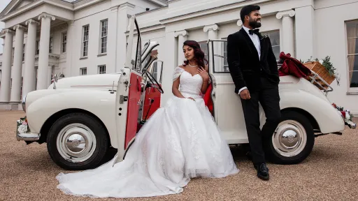 Bride and groom standing beside cream 1963 Morris Minor Convertible with red interior at wedding venue entrance.