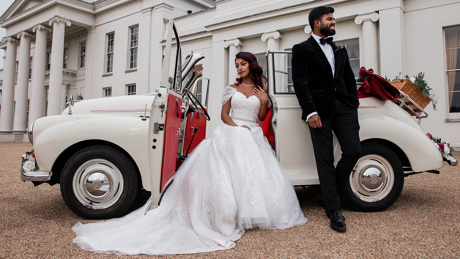 Bride and groom standing beside cream 1963 Morris Minor Convertible with red interior at wedding venue entrance.