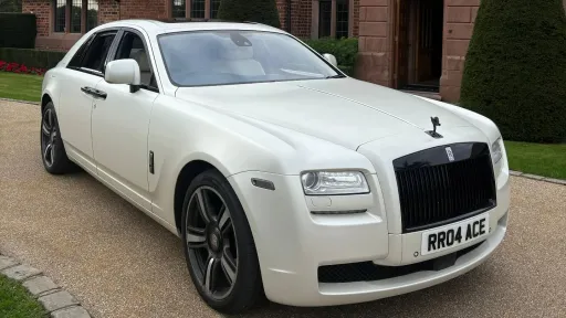 White Rolls-Royce Ghost Black Badge photographed from the front three-quarter angle, parked on a light gravel driveway with greenery behind.