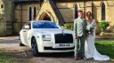 White Rolls-Royce Ghost parked at the front of a stone church, front three-quarter view, with bride and groom standing beside the car on a sunny day.