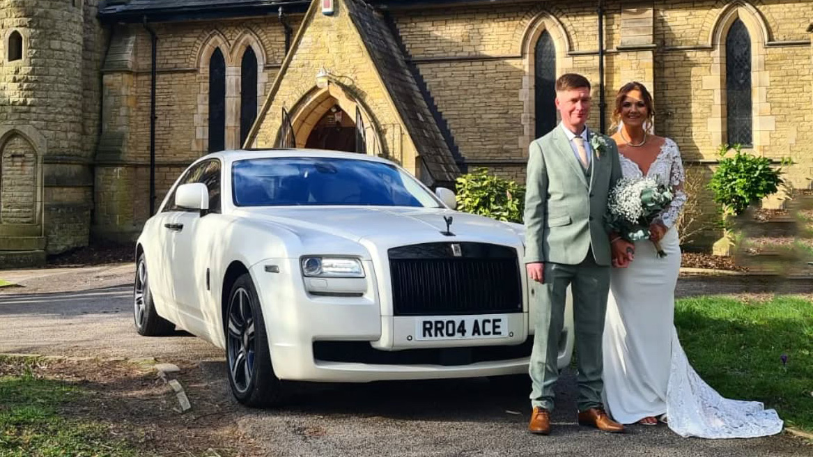 White Rolls-Royce Ghost parked at the front of a stone church, front three-quarter view, with bride and groom standing beside the car on a sunny day.