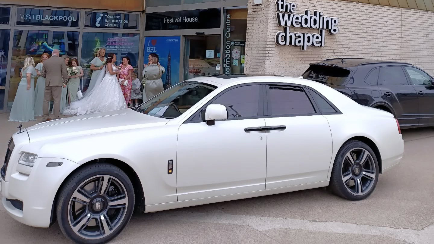 White Rolls-Royce Ghost shown in full left side profile, parked outside a modern wedding chapel with guests gathered near the entrance.