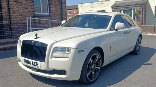 White Rolls-Royce Ghost Black Badge parked on a paved driveway, front three-quarter angle, with brick buildings in the background.