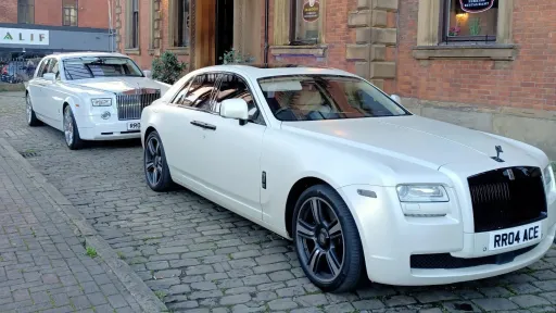 Two white Rolls-Royce cars parked in a line on a cobbled courtyard, front car in three-quarter view, red brick venue building behind.