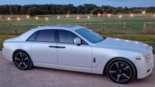 White Rolls-Royce Ghost Black Badge in full side profile, parked on gravel at dusk, countryside field and string lights in the background.
