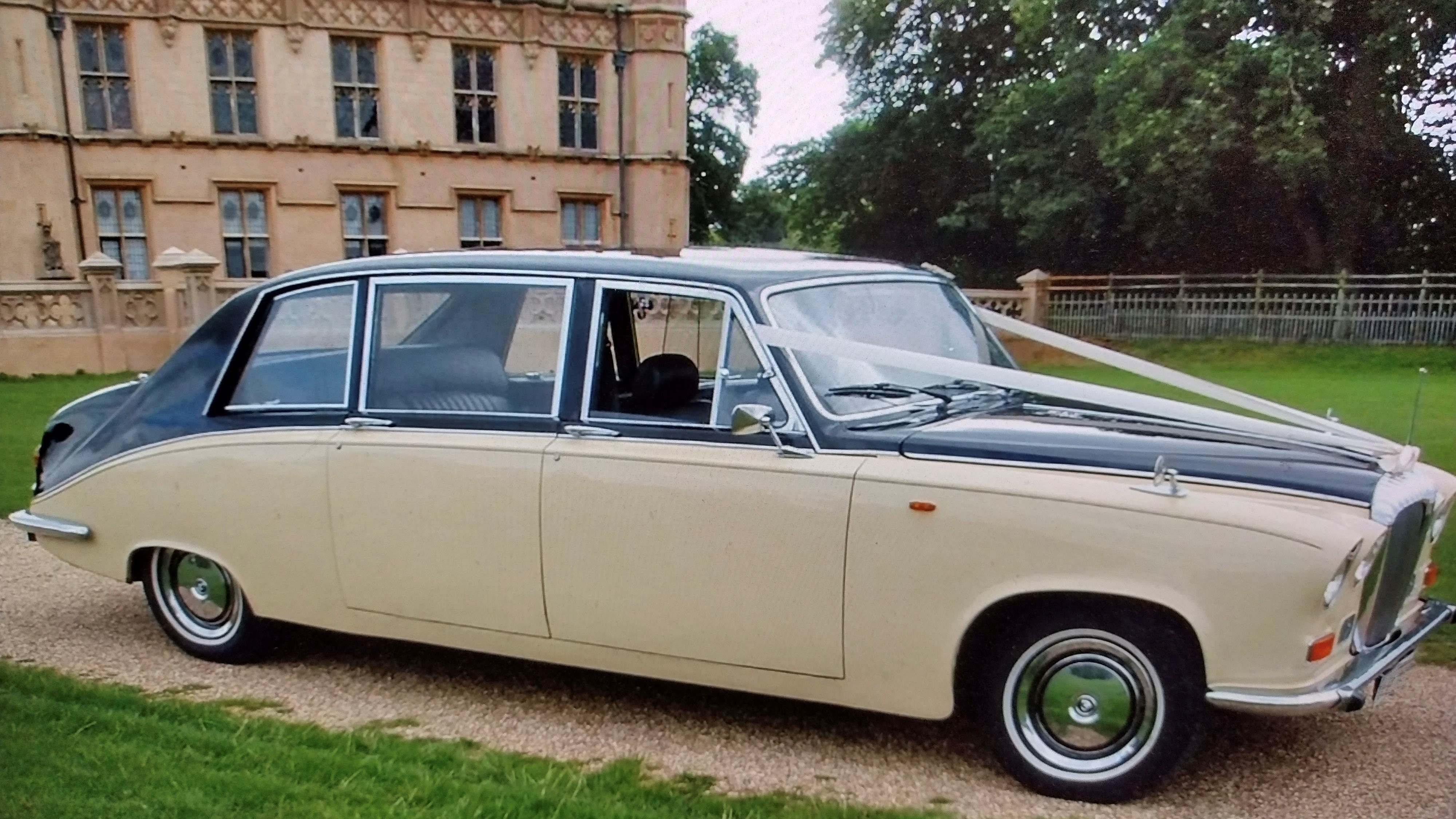 Black and ivory Daimler limousine parked outside a grand stately building, angled front side view showing chrome grille and long body.