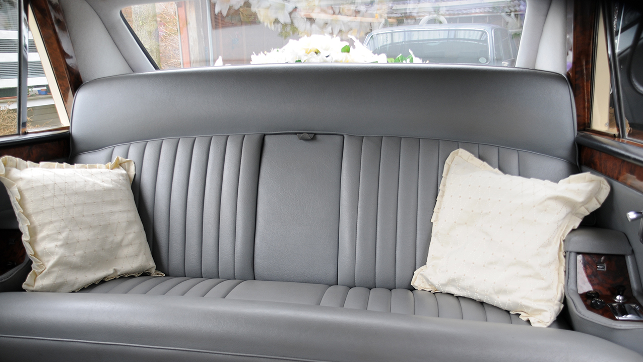 Rear cabin interior of classic Daimler limousine with silver leather bench seating, pleated upholstery and cream cushions.