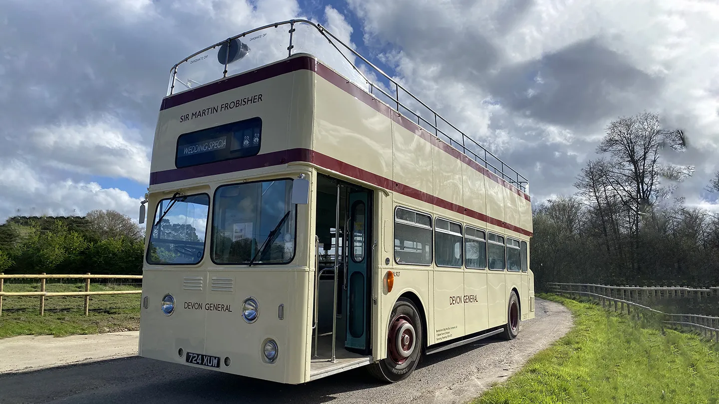 Cream vintage open top Leyland double decker bus driving along a country lane under cloudy skies.