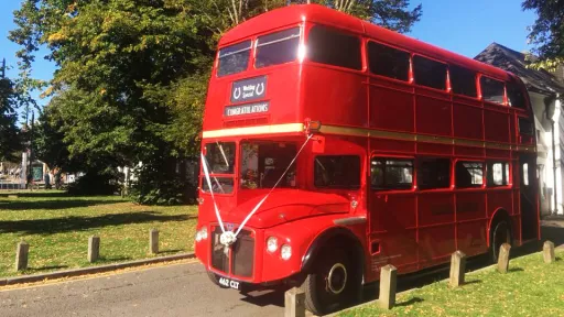Red AEC Routemaster double decker wedding bus parked on a grass area beside a country wedding venue with trees and blue sky.