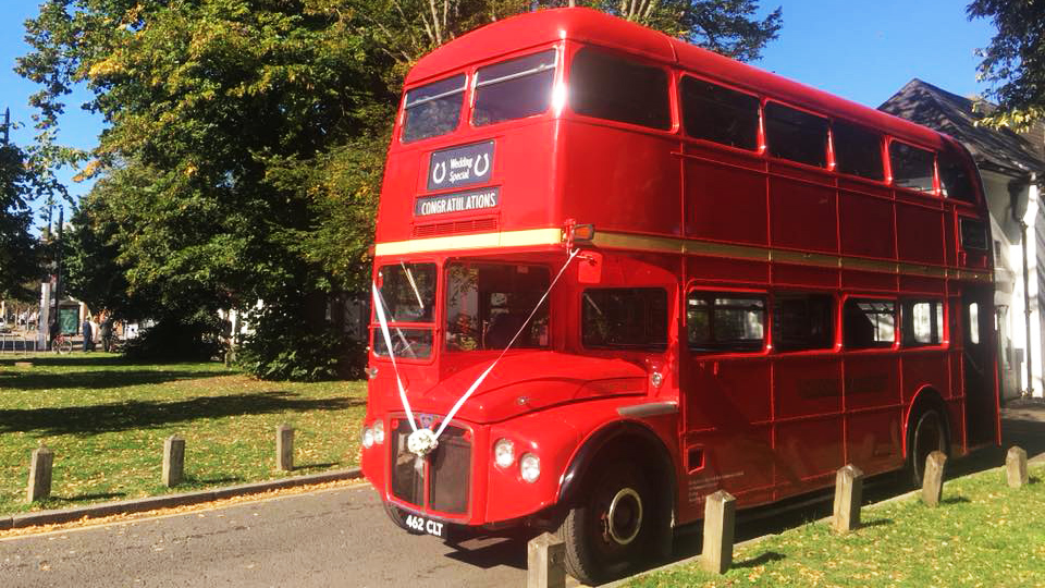 Red AEC Routemaster double decker wedding bus parked on a grass area beside a country wedding venue with trees and blue sky.
