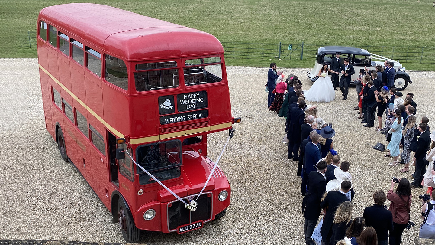 Aerial view of a red vintage Routemaster double decker bus decorated with white wedding ribbons surrounded by guests outside a country venue.