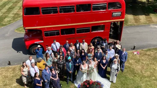 Aerial view of wedding guests gathered on grass with a red Routemaster double decker bus parked behind them.