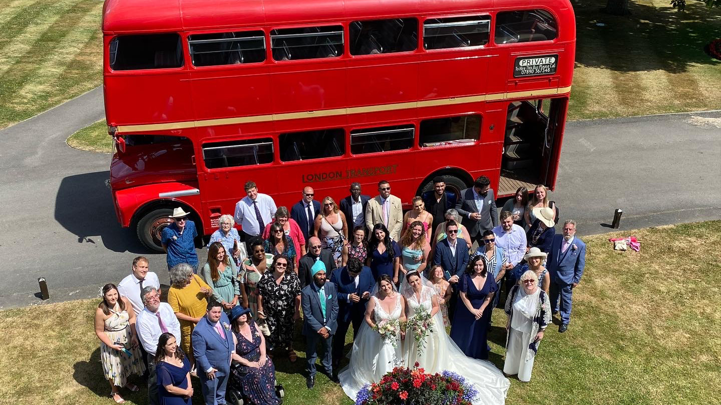 Aerial view of wedding guests gathered on grass with a red Routemaster double decker bus parked behind them.