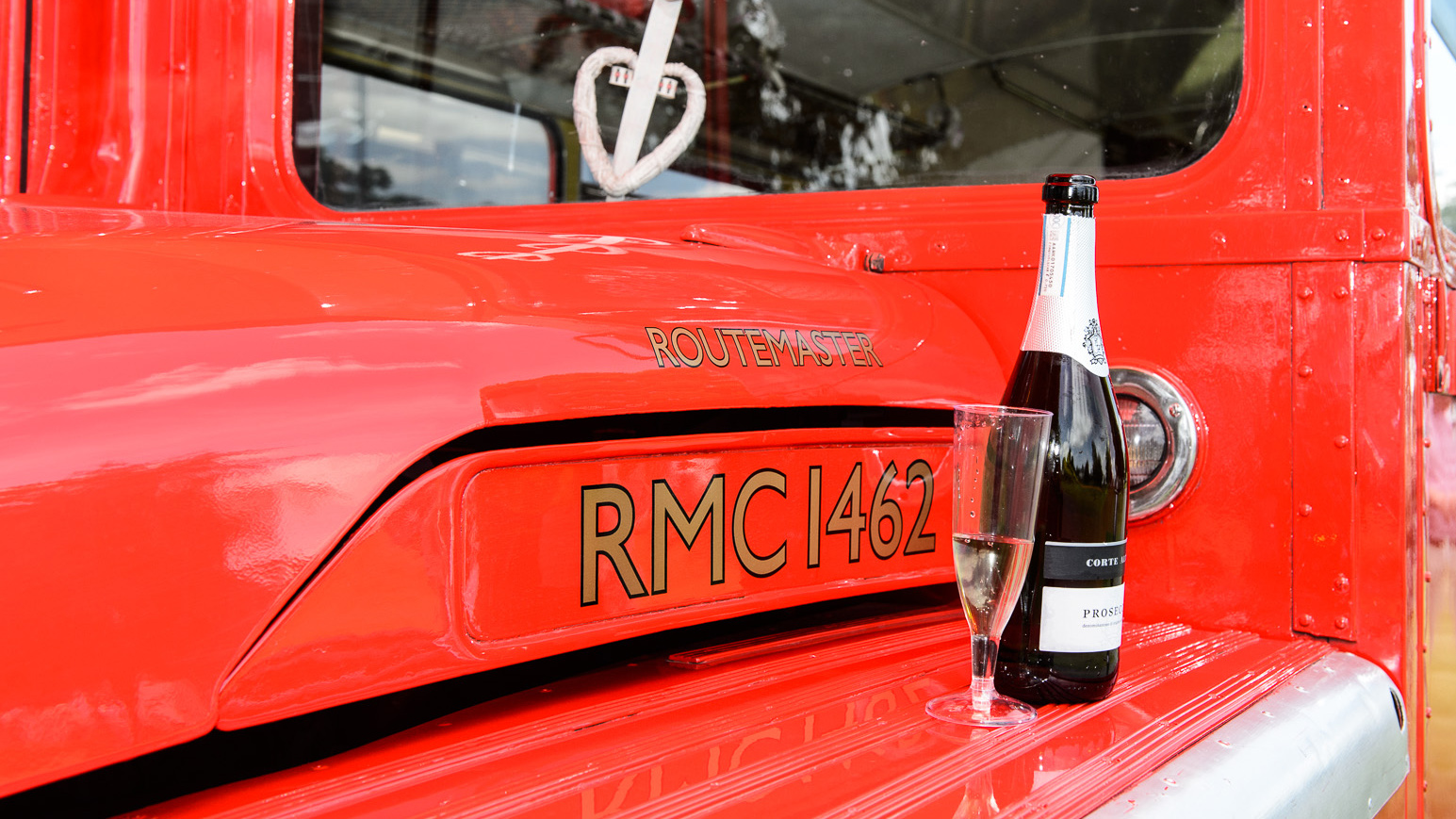 Bottle of champagne placed on the rear ledge of a red AEC Routemaster bus with wedding ribbons visible on the vehicle.