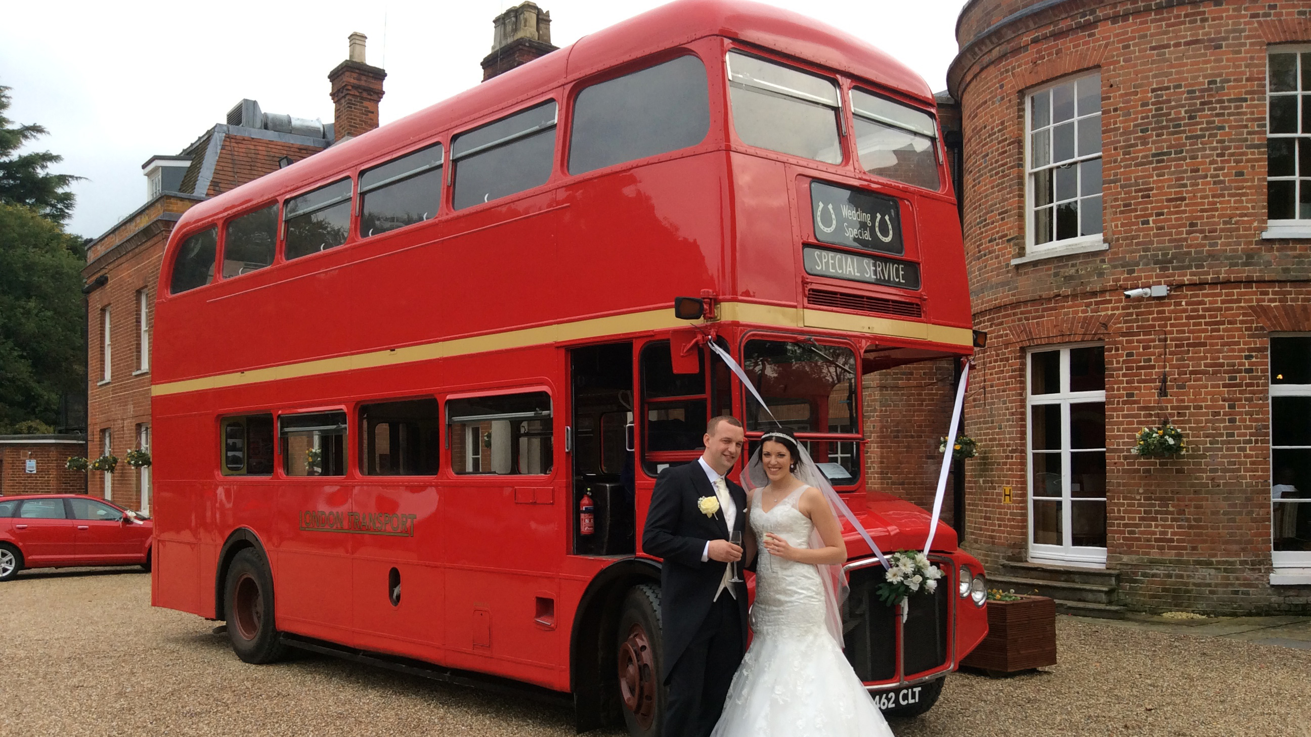 Bride and groom standing beside a red vintage Routemaster double decker bus outside a brick wedding venue.