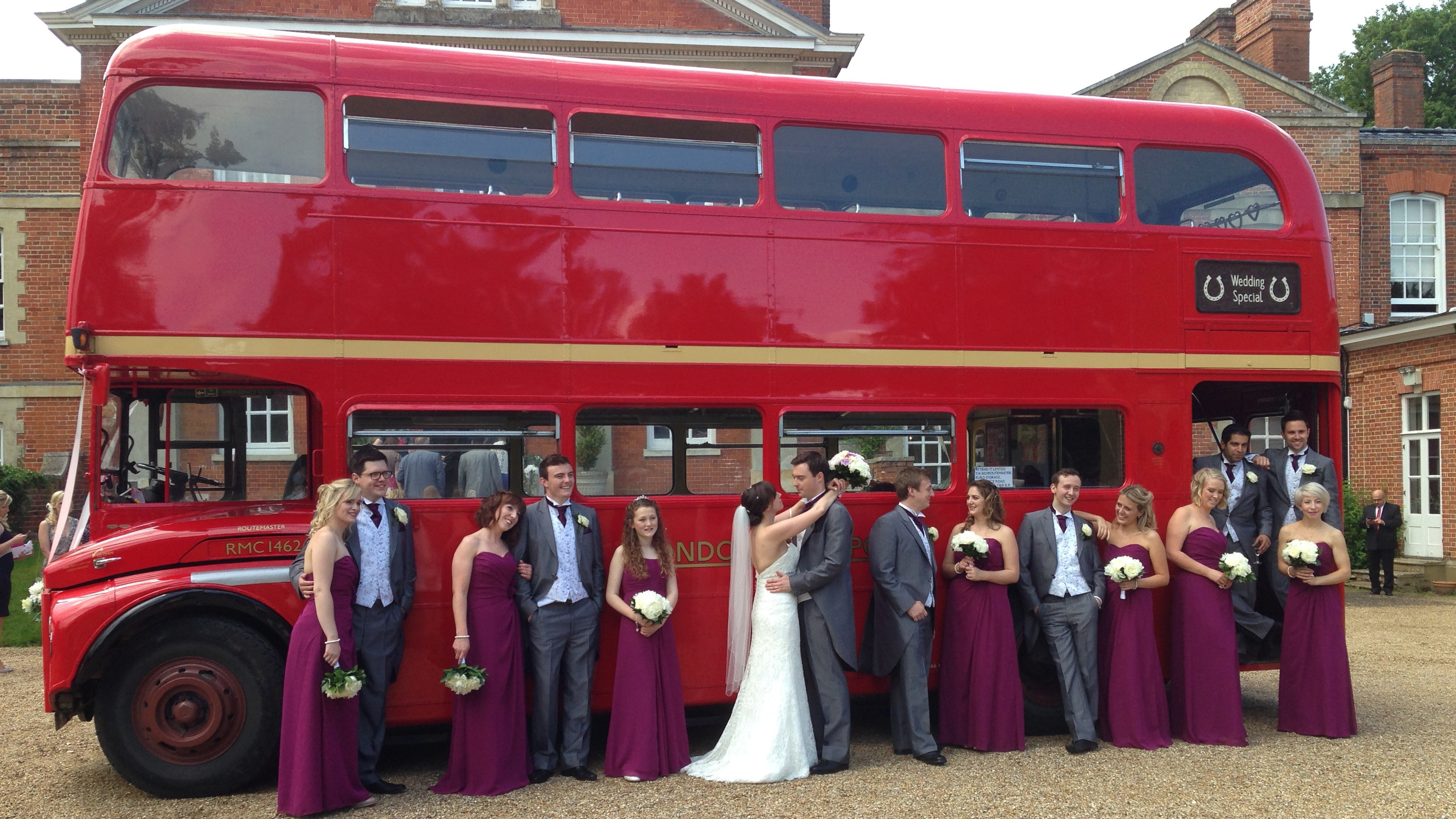 Bride and bridesmaids posing beside a red Routemaster double decker wedding bus outside a historic venue.