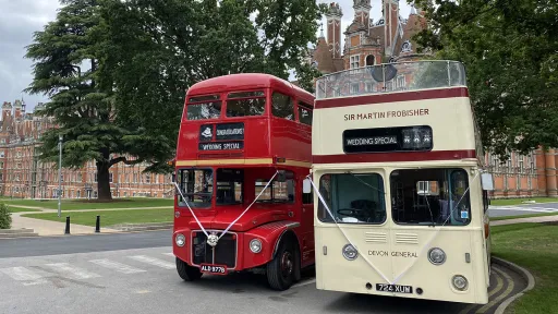 Cream Leyland open top double decker bus parked beside a red Routemaster bus outside a historic venue.