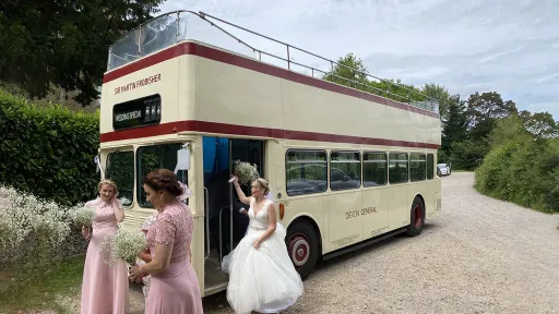 Bride and bridesmaids standing beside a cream Leyland double decker wedding bus outside a countryside venue.