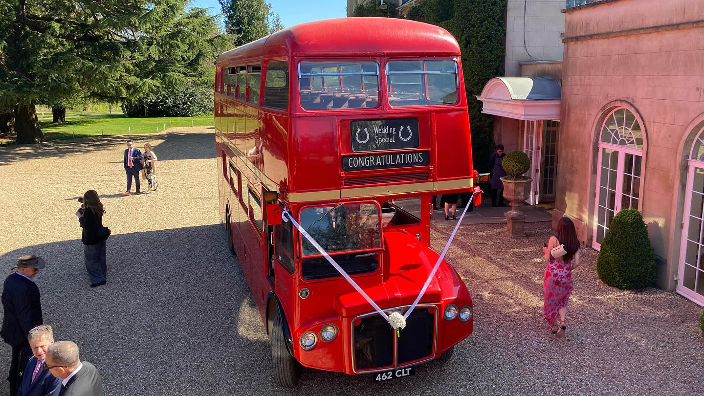 Front view of a red Routemaster double decker bus decorated with white wedding ribbons parked in a courtyard.