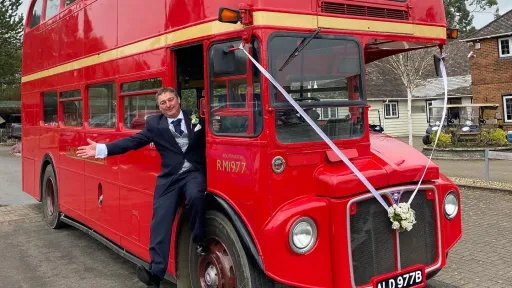 Groom standing on the step of a red vintage Routemaster double decker wedding bus decorated with white ribbons.