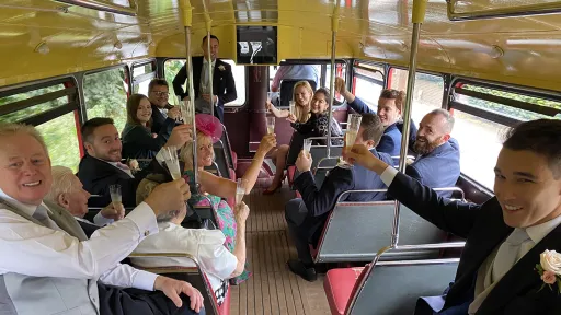 Wedding guests seated inside a vintage red Routemaster double decker bus raising glasses during the journey.