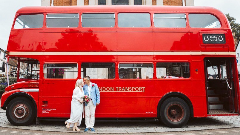 Side view of a red Routemaster double decker bus showing the open rear platform with bride and groom nearby.