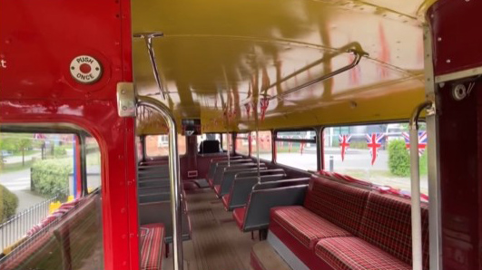 Lower deck interior of a vintage Routemaster bus with traditional bench seating and large windows.
