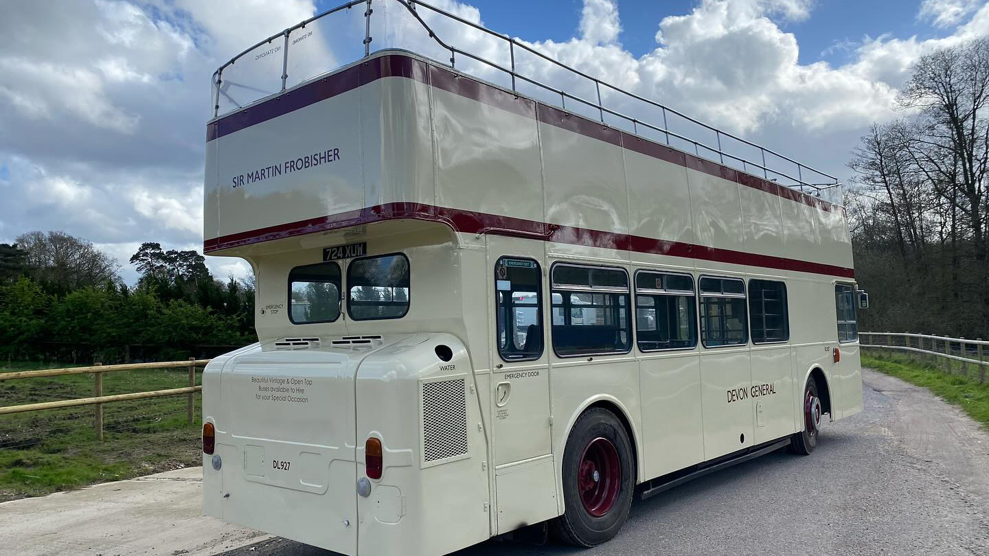 Rear view of a cream open top Leyland double decker bus driving along a rural road.