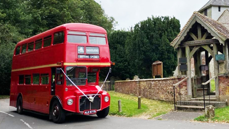 Red vintage Routemaster double decker wedding bus parked outside a small countryside chapel.
