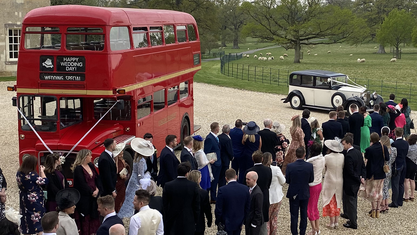 Red Routemaster double decker bus with wedding guests gathered nearby and a vintage car parked in the background.