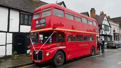 Red Routemaster double decker bus decorated with white wedding ribbons parked beside historic buildings.