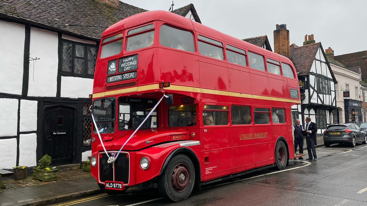 Red Routemaster double decker bus decorated with white wedding ribbons parked beside historic buildings.