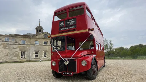 Front view of a red vintage Routemaster wedding bus decorated with white ribbons parked in front of a stately venue.