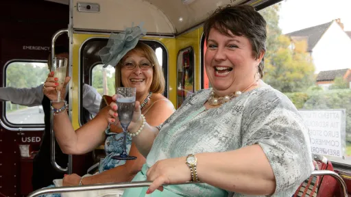 Wedding guests smiling and holding drinks while seated inside a vintage red Routemaster wedding bus.