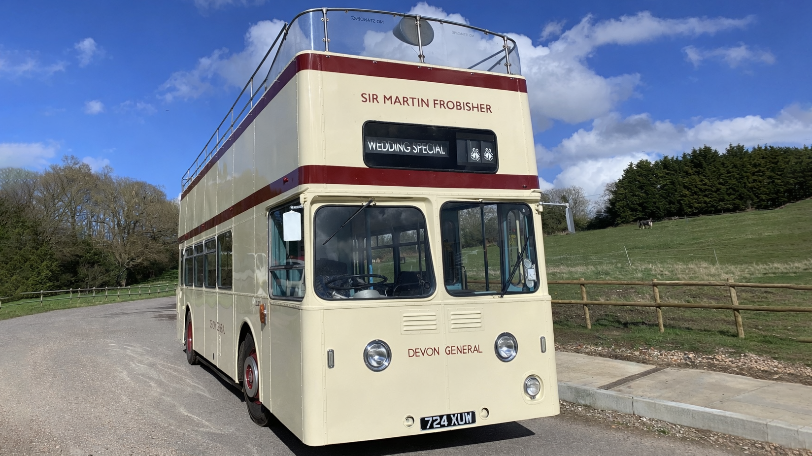 Front view of a cream open top Leyland double decker wedding bus parked beside countryside fields.