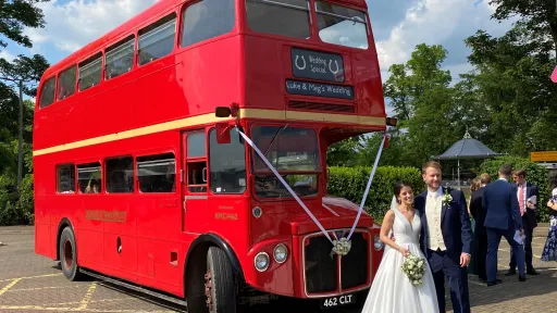 Bride and groom posing beside a red AEC Routemaster double decker wedding bus decorated with white ribbons.