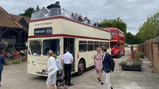 Wedding guests gathered beside a cream vintage Leyland double decker bus outside a countryside venue.
