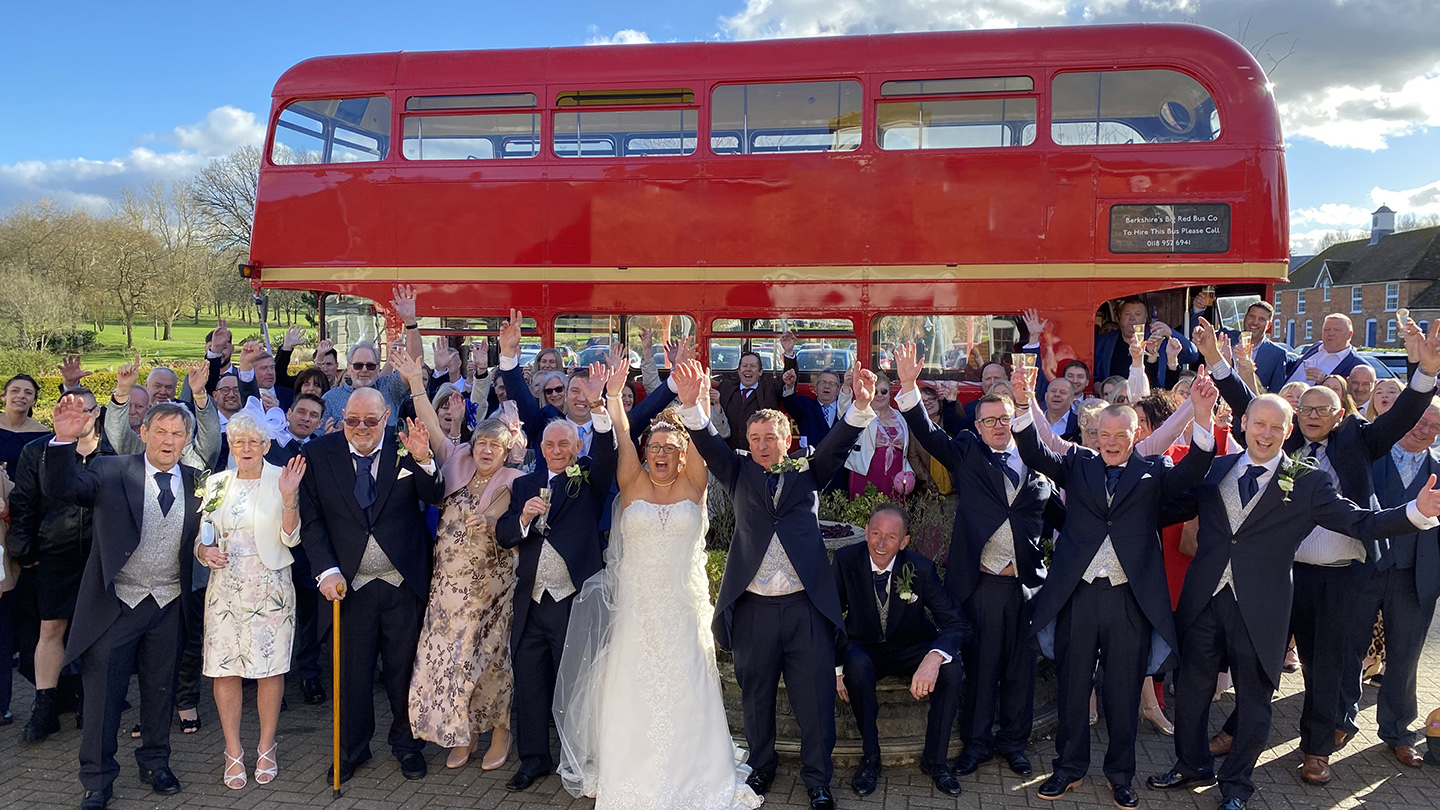 Large wedding group posing for photos with a red Routemaster double decker bus behind them.